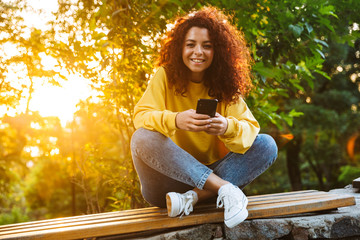 Cheerful smiling cute young student curly girl sitting on bench outdoors in nature park with...
