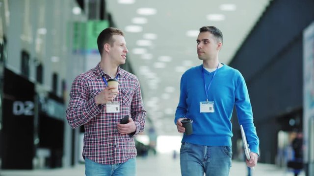 Two young businessmen with id cards walking down office lobby and talking over coffee. Men holding cell phone and laptop computer. Business event attendees networking at coffee break