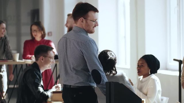 Happy young CEO businessman entering comfortable modern multiethnic loft office, sitting down at table smiling.