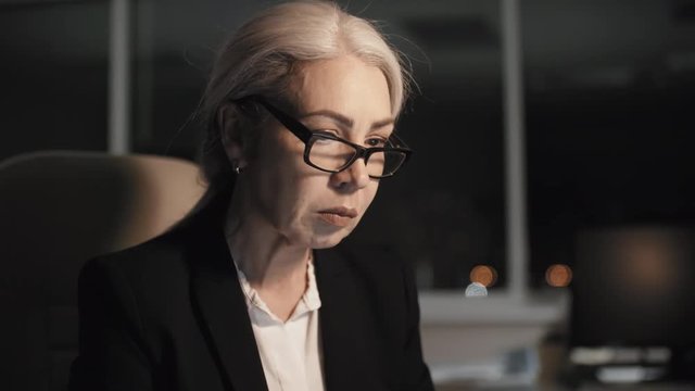 Close-up face shot of grey-haired Caucasian career woman, wearing formal black suit and white blouse, working in office late at night, looking at screen with her lips moving, and straightening glasses