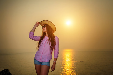 Beautiful long-haired woman against sunset on the sea. Young girl in sunglasses holding a hat and standing by the water. Warm evening on the coast, soft sunset light, glare on calm water.