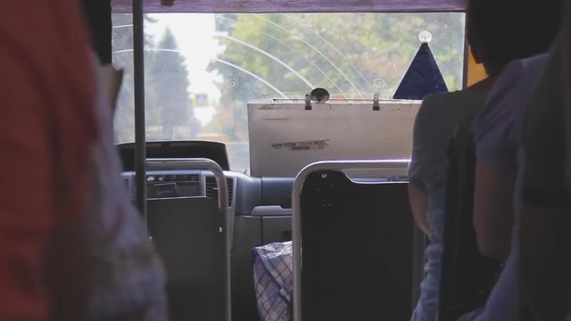 Inside view of a regular bus. The landscape changes behind the windshield. People are sitting in the passenger seats. Passage between the seats. Summer sunny day. Public transport