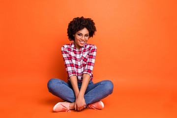 Full length body size photo beautiful nice she her dark skin lady sitting floor legs crossed amazing look wear specs casual jeans denim checkered plaid shirt isolated bright orange background