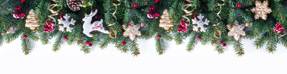 Festive Christmas border - green fir branches decorated with ginger Christmas glazed cookies in form of reindeer and snowflakes, red berries, banner format,  isolated on white.