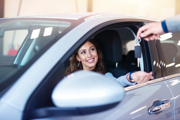 Car dealership. An attractive brunette woman sitting in her brand new car and taking keys from vehicle dealer.