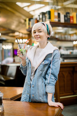 Pretty smiling blonde girl in jeans clothes, holding glass with cocktail in hands. Indoor portrait of joyful young woman in casual clothes laughing in cafe, enjoying cold cocktail in warm day.