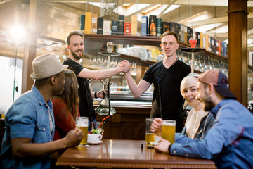 Happy multiethnical friends having drinks in a bar, they are sitting at a wooden table with beers. Two guys holding hookah