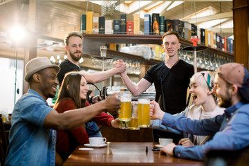 Happy multiethnical friends having drinks in a bar, they are sitting at a wooden table with beers. Two guys holding hookah