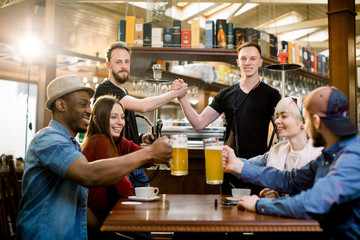 Happy multiethnical friends having drinks in a bar, they are sitting at a wooden table with beers. Two guys holding hookah