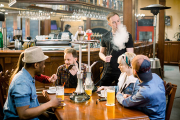 Large group multiracial friends boys and girls sitting at table in cafe, drinking beer and cocktails. Young guy smokes hookah