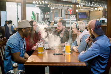 Young multiracial people vapor hookah pipe in shisha bar lounge. Women and men friends relax in cafe indoor. Celebration, party, proposal, birthday. Bad habits, party, addiction.