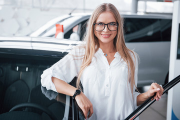 It's her birthday gift. Cute girl in eyewear stands near the car in auto saloon. Probably her next purchase