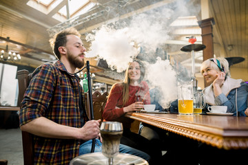 Close-up of Young man in casual wear smoking traditional hookah pipe. man exhaling smoke. Friends in cafe having fun