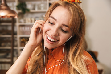 Fototapeta premium Happy optimistic young blonde girl in orange shirt at the kitchen listening music with earphones.