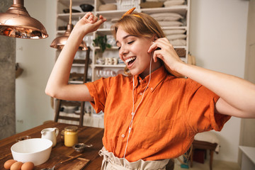 Happy optimistic young blonde girl in orange shirt at the kitchen listening music with earphones.