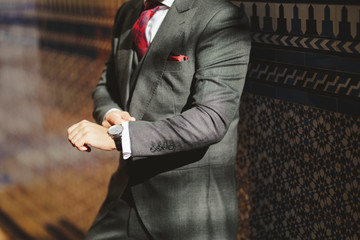 Unrecognizable man in a suit, closing his watch while leaning against the wall.