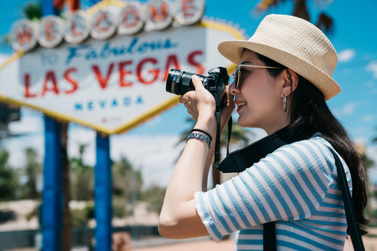 Portrait of beautiful laughing asian chinese girl wearing blue striped t-shirt making photos at summer travel trip. woman photographer taking pictures by camera while standing by welcome sign in vegas