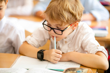 Naklejka na ściany i meble Blonde boy with big black glasses sitting in classroom, studing, smiling. Education on elementary school, first day at school