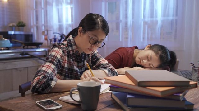 young asian college girl student in glasses sitting at desk with books near tired friend. hard working lady concentrated doing homework at night. female touching roommate shoulder waking her up.