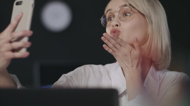 Cheerful woman sending air kiss for mobile selfie in night office