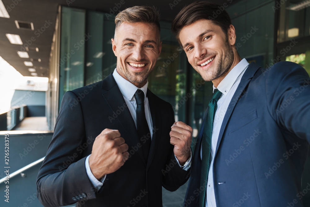 Wall mural photo of beautiful two businessmen smiling at camera and taking selfie while standing outside job ce