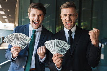 Image of excited businessmen partners rejoicing and holding bunch of cash money while standing...