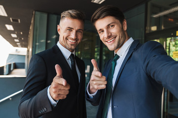 Photo of beautiful two businessmen dressed in formal suit showing thumbs up and taking selfie while...