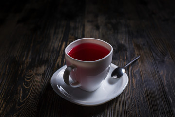 A cup of red berry or karkade tea in white cup with teaspoon on a dark wooden table. Concept for menu.