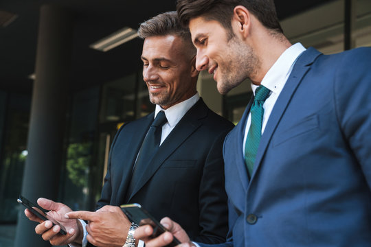 Image of happy businessmen partners standing outside office center and holding cellphones together during working meeting