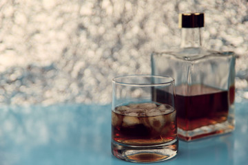 A glass of whiskey with ice and a square transparent bottle on a light blue background and silver bokeh. Free space on the left, horizontal, close-up, blur. Alcohol culture concept.