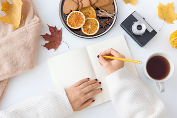 technology and season concept - woman's hands writing to notebook with pencil, autumn leaves, cup of tea, gingerbread cookies with dried orange slices and film camera on white background