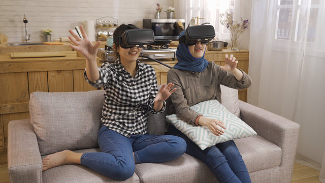 two asian best girl friends trying on VR headsets sitting on couch having fun at home. japanese and islam women playing games with virtual reality goggles relax on sofa in kitchen on weekend.