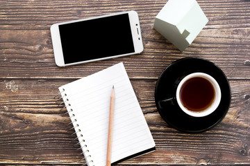 Mobile phone; blank spiral notebook; pencil; coffee cup and house model on wooden desk