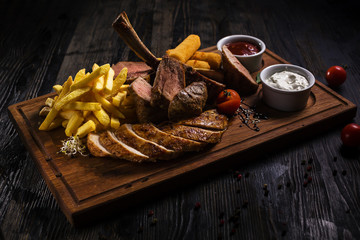 Delicious ribs with french fries and chiken and fries cheese sticks with sauce on wooden cutting board on wooden table in low key