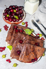 Chocolate, thin round pancakes dressed with cherry jam. Near in a transparent bowl are fresh cherries, on a gray background. There are also green gooseberries and red currants. 