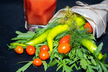 A bunch of vegetables. It consists of hot pepper, dill, arugula, cherry tomatoes, all of which are folded into paper, against a black background. Healthy food. in the background is tomato juice 