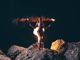 Fototapety  Young sexy woman with a gorgeous slim body in bikini is staying (posing) at the dark night on the rock with the lit by fire torches in her hands. Black sky at the background