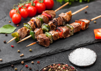 Grilled pork and chicken kebab with paprika on stone chopping board with salt, pepper and tomatoes on black background.