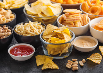 All classic potato snacks with peanuts, popcorn and onion rings and salted pretzels in bowl plates on black background. Twirls with sticks and potato chips and crisps with nachos and cheese balls.