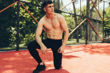 Outdoors image of muscular young man streching out against nature background. Athlete man looking away, stretching his leg and listening the music on earphones. Sport, lifestyle and people concept.