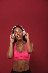 Pleased strong young african sports woman posing isolated over red wall background listening music with headphones.