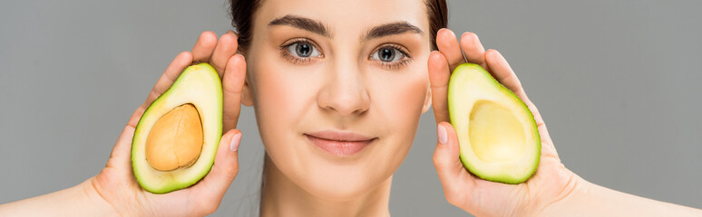 panoramic shot of young woman holding avocado halves isolated on grey