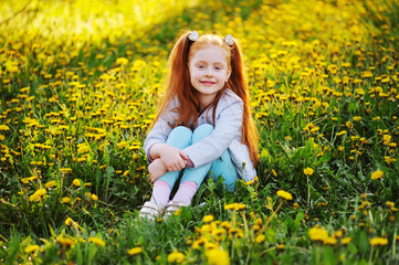 Naklejka na ściany i meble portrait of a child - a little cute red-haired girl on the background of a field of dandelions and greenery.