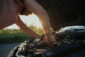 A young man repairs a car at sunset outside the city