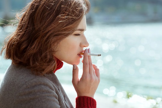 beautiful stylish sad stressful young woman in sweater and coat smoking cigarette near the river