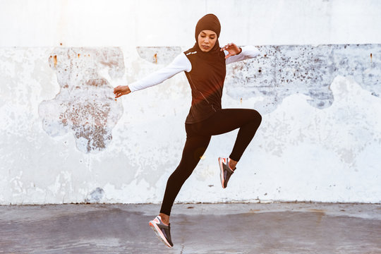 Concentrated Muslim Sports Fitness Woman Dressed In Hijab And Dark Clothes Posing Make Sport Stretching Exercises Outdoors At The Street Running.