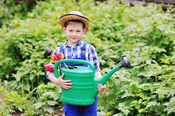 child boy in a straw hat in a blue work suit gardener with a bouquet of tulips and a green watering can in his hands smiling at the background of the garden