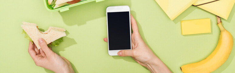 cropped view of woman holding smartphone and sandwich in hands near lunch box with food, banana and stationery