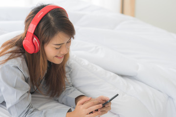 Young happy Asian woman lie down on bed playing smart tablet and listening music in modern bedroom