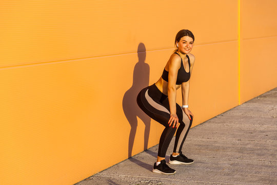 Young Attractive Girl In Black Sporwear Practicing Sport Exercise On Street, Standing On Squat Near Orange Wall Background, Take Rest After Hard Workout, Outdoor, Looking At Camera